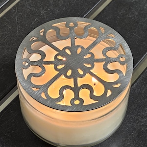 May include: A lit candle in a clear glass jar with a decorative silver metal lid. The lid features an openwork design. The candle is on a dark surface.