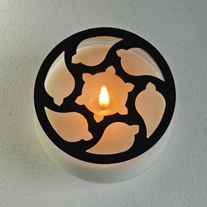 May include: A lit candle in a white container with a black, decorative, leaf-shaped cutout design. The candle flame glows brightly, illuminating the creamy wax within the container. The design is circular.