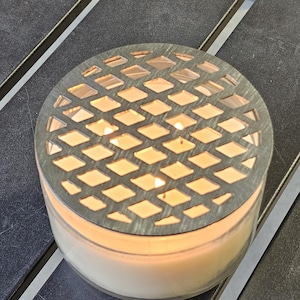 May include: A lit candle in a clear glass jar with a decorative metal lid. The lid has a diamond-shaped pattern. The candle is on a dark surface.