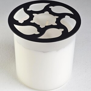 May include: A white candle in a clear glass jar with a black decorative lid. The lid has a circular design with leaf and turtle-shaped cutouts. The candle has a visible wick.