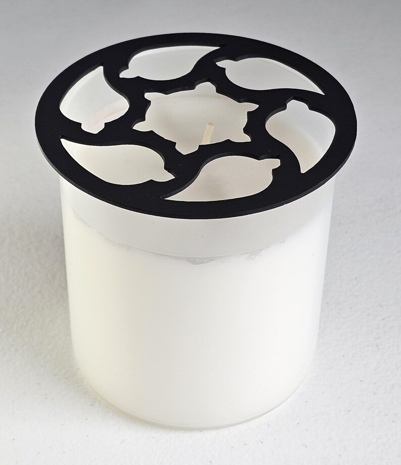 May include: A white candle in a clear glass jar with a black decorative lid. The lid has a circular design with leaf and turtle-shaped cutouts. The candle has a visible wick.