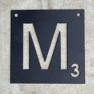 May include: Black metal square sign with a white cutout of the letter M and the number 3.