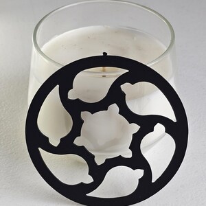 May include: A clear glass candle holder with a white candle inside. A black metal candle topper with a decorative cutout design is placed on top of the candle. The candle topper has a circular shape with a floral pattern.
