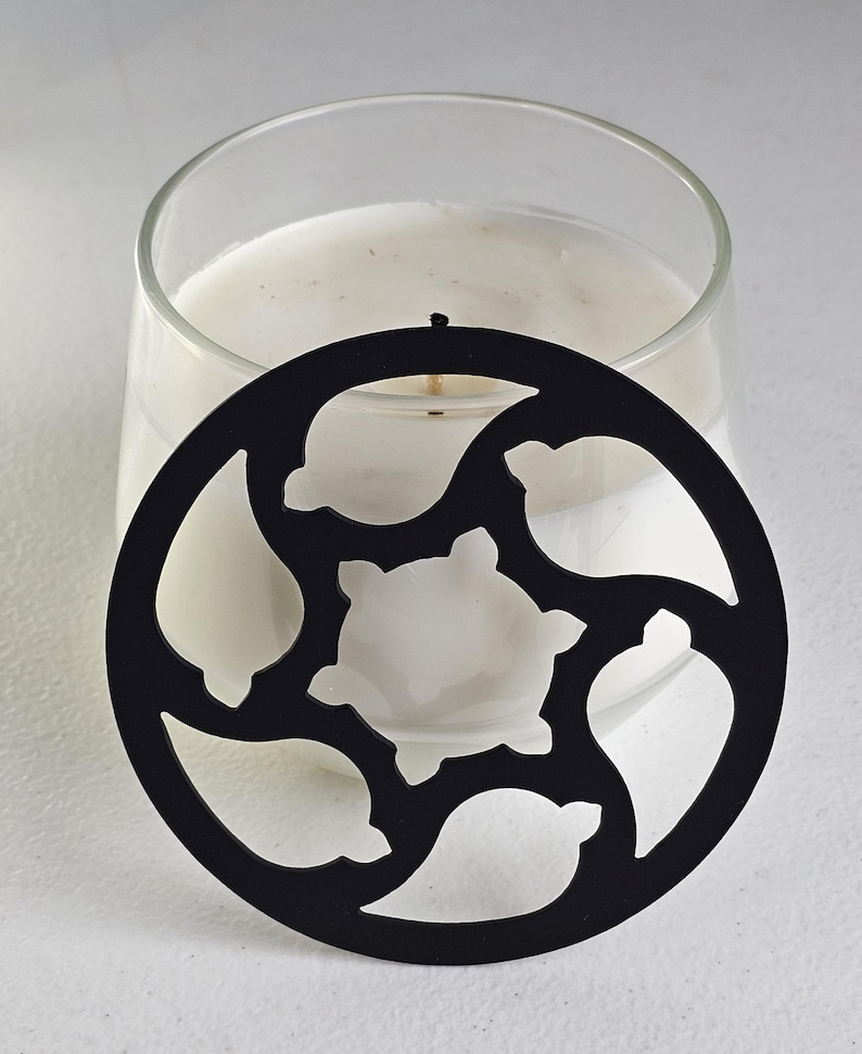May include: A clear glass candle holder with a white candle inside. A black metal candle topper with a decorative cutout design is placed on top of the candle. The candle topper has a circular shape with a floral pattern.