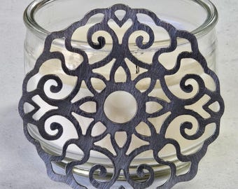 May include: A decorative, circular metal candle lid with an intricate, openwork design. The lid is dark gray and sits atop a clear glass candle jar. The candle inside is white, and the overall aesthetic is elegant and ornate.