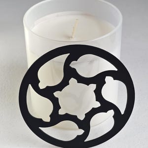 May include: A white candle in a clear glass jar with a black decorative lid. The lid has a circular shape with cut-out leaf and star-like designs. The candle has a single wick.