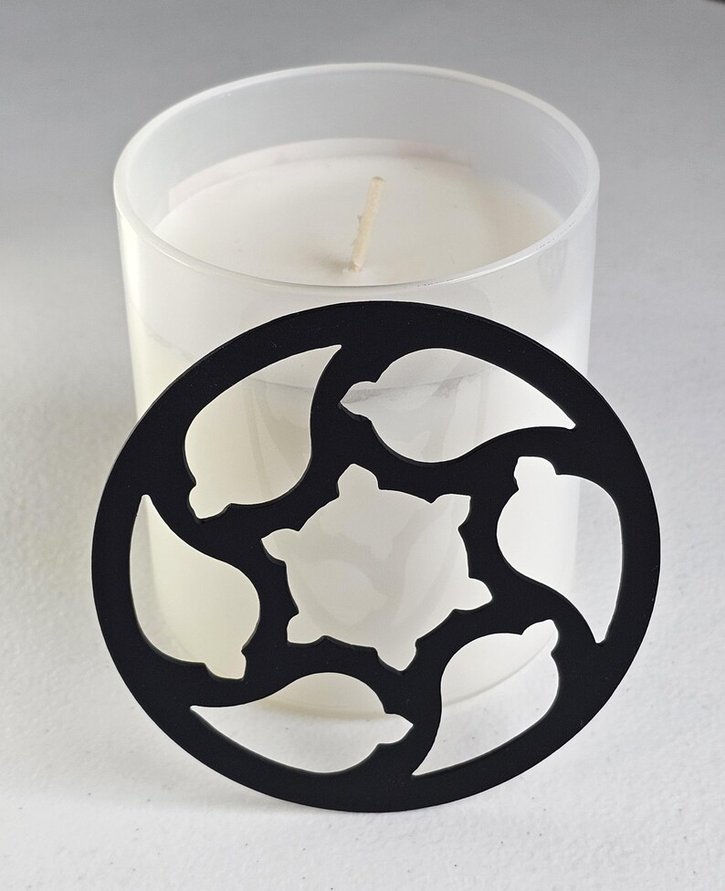 May include: A white candle in a clear glass jar with a black decorative lid. The lid has a circular shape with cut-out leaf and star-like designs. The candle has a single wick.