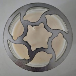 May include: Overhead view of a candle in a glass jar, with a decorative metal lid. The lid has leaf-shaped cutouts around the edge and a central star-shaped cutout. The candle wax is a creamy beige color.
