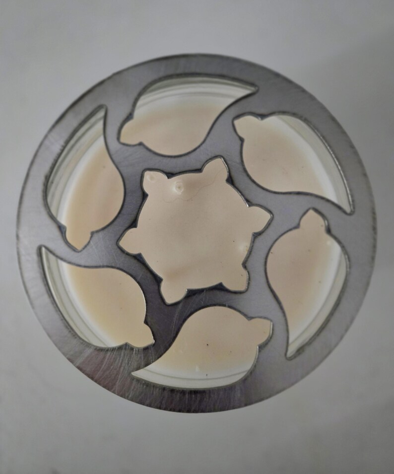 May include: Overhead view of a candle in a glass jar, with a decorative metal lid. The lid has leaf-shaped cutouts around the edge and a central star-shaped cutout. The candle wax is a creamy beige color.