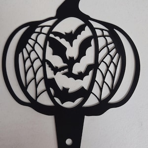 May include: Black metal Halloween pumpkin decoration with a carved face, bats, and spiderwebs. The pumpkin has a stem and a stake for easy placement in a garden or yard. A festive accent for Halloween.