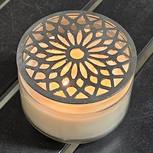May include: A burning candle in a clear glass jar, topped with a decorative metal lid featuring a floral design. The candle wax is a creamy hue, and the flame is visible through the lid's cut-out pattern. The candle is on a dark surface.