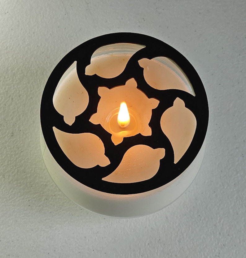 May include: A lit candle in a white container with a black, decorative, leaf-shaped cutout design. The candle flame glows brightly, illuminating the creamy wax within the container. The design is circular.