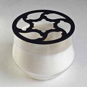 May include: A clear glass candle holder filled with white wax, topped with a black decorative lid. The lid has a floral design with cut-out shapes. The candle holder sits on a white surface.