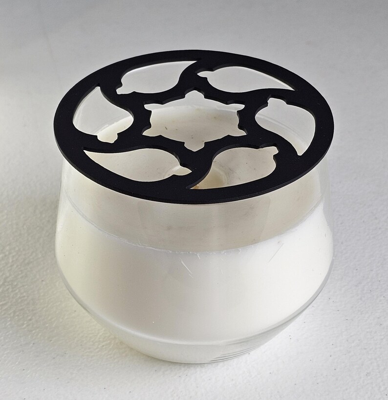 May include: A clear glass candle holder filled with white wax, topped with a black decorative lid. The lid has a floral design with cut-out shapes. The candle holder sits on a white surface.
