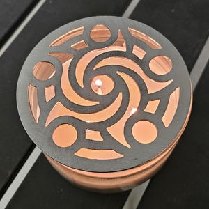 May include: A lit candle in a copper-colored container, topped with a decorative silver metal lid. The lid features a swirling, openwork design. The candle's flame is visible through the lid's pattern, casting a warm glow.