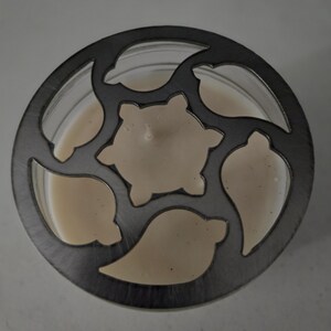 May include: A candle in a clear glass jar with a decorative metal lid. The lid features a pattern of stylized leaves and a central star shape. The candle wax is a creamy off-white color.