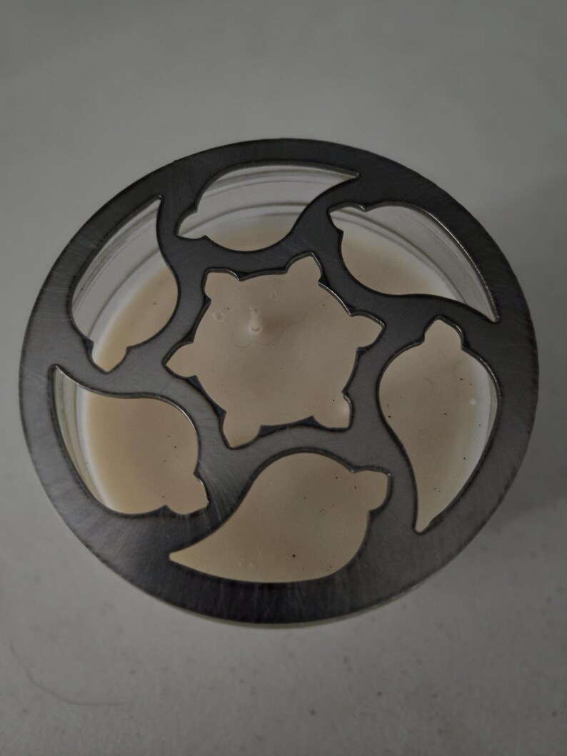 May include: A candle in a clear glass jar with a decorative metal lid. The lid features a pattern of stylized leaves and a central star shape. The candle wax is a creamy off-white color.
