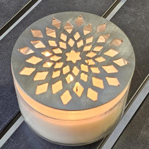 May include: A lit candle in a clear glass jar with a decorative metal lid. The lid has a pattern of diamond and star-shaped cutouts. The candle is on a dark surface.