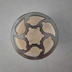 May include: A circular metal cookie cutter with a floral design. The cutter has a series of leaf-shaped cutouts around the edge and a six-petal flower shape in the center. The cutter is filled with a light beige dough.