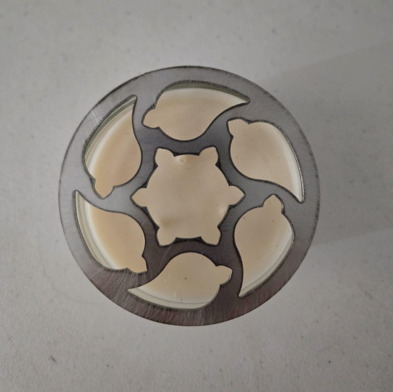 May include: A circular metal cookie cutter with a floral design. The cutter has a series of leaf-shaped cutouts around the edge and a six-petal flower shape in the center. The cutter is filled with a light beige dough.