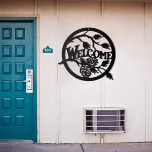 May include: Black metal welcome sign with pine cones and branches in a circle. The sign is mounted on a white wall next to a blue door.