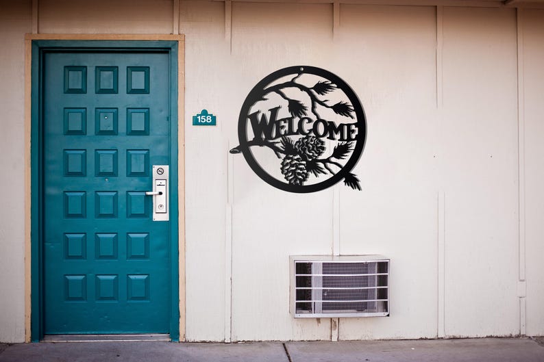 May include: Black metal welcome sign with pine cones and branches in a circle. The sign is mounted on a white wall next to a blue door.