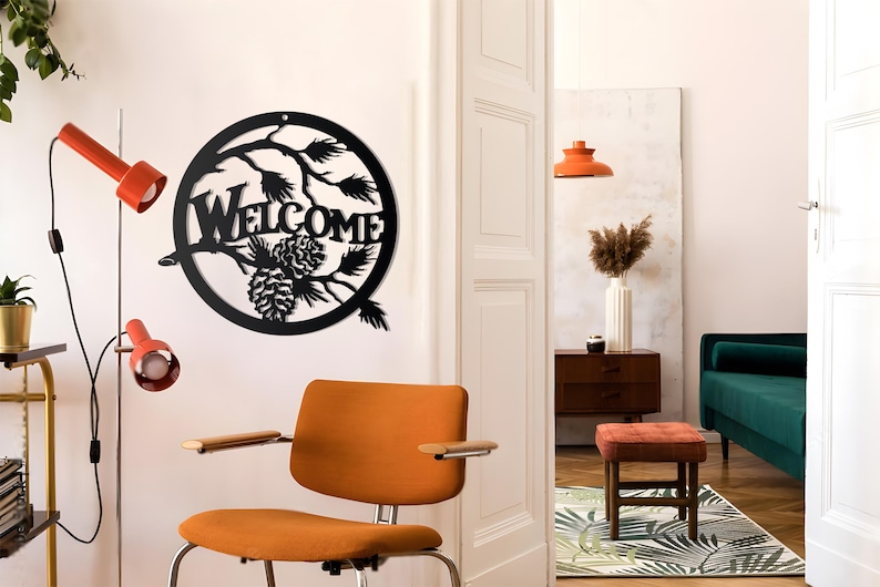 May include: Black metal welcome sign with pine cones and branches in a circle. The sign is hanging on a white wall in a living room with a brown chair and a green sofa.