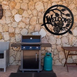 May include: Black metal welcome sign with a pine cone and branches design, hanging on a stone wall. A black gas grill with a propane tank is in the foreground.
