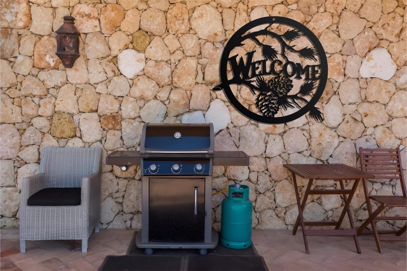 May include: Black metal welcome sign with a pine cone and branches design, hanging on a stone wall. A black gas grill with a propane tank is in the foreground.