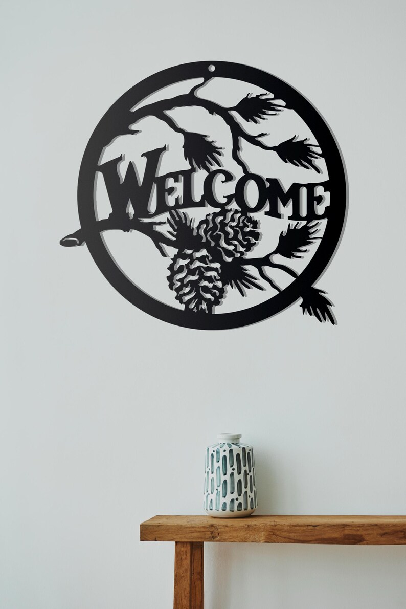 May include: Black metal welcome sign featuring pine branches and pine cones. The sign is round and has a rustic feel.