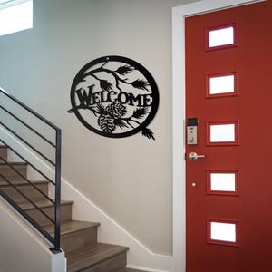 May include: Black metal welcome sign featuring a pine cone and branch design. The sign is hanging on a white wall next to a red door with glass panels.