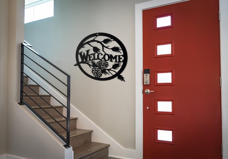 May include: Black metal welcome sign featuring a pine cone and branch design. The sign is hanging on a white wall next to a red door with glass panels.