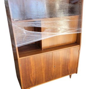 May include: A mid-century modern wooden cabinet with a warm brown finish. The cabinet features a combination of open shelving and closed storage with two doors. The unit is wrapped in clear plastic for protection.