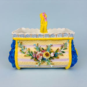 May include: A rectangular ceramic basket with a yellow and lavender striped pattern, featuring a central floral arrangement of sculpted roses and leaves. The basket has a yellow handle, blue side accents, and a scalloped rim with blue trim.