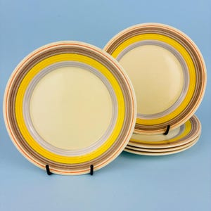 May include: Set of four cream-colored ceramic plates with concentric rings of yellow, gray, brown, and red. The plates are stacked and displayed on a light blue background, showcasing a retro design.