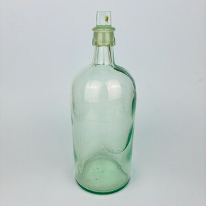 May include: A clear, light green glass bottle with a rounded body and a narrow neck. The bottle features a thick, cylindrical glass stopper inserted into the top opening. The glass has a smooth, translucent finish with visible air bubbles.