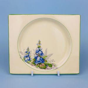 May include: Square cream plate with a green border and a central circular design. It features a floral motif of blue bell-shaped flowers and green leaves. The plate is displayed on a white stand against a blue background.
