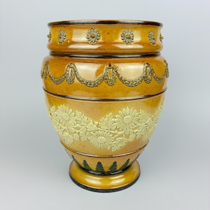 May include: A glazed mustard-yellow ceramic vase featuring a raised relief pattern of cream-coloured daisies and leaves around the centre, a decorative garland border near the top, a row of small flower motifs at the rim, and dark leaf accents at the base.