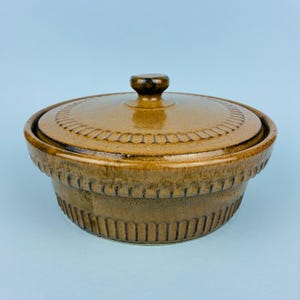 以下が含まれることがあります： 蓋付きの丸い茶色の陶器製キャセロール皿。皿の外側と蓋の縁には、縦方向の畝模様が繰り返し施されています。釉薬は斑点のある光沢仕上げで、濃い茶色の色調が特徴です。