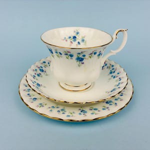 Peut inclure: Un ensemble tasse et soucoupe en porcelaine blanche orné d'un motif délicat de petites fleurs bleues et violettes avec des feuilles vertes. Les bords de la tasse, de la soucoupe et de l'assiette sont finis par un fin liseré doré festonné.