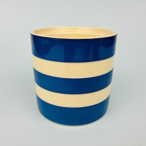 May include: A cylindrical ceramic vessel featuring a repeating horizontal striped pattern in deep navy blue and cream. The container has a smooth, glossy finish and an open top, with the interior rim showing a light cream colour consistent with the stripes.