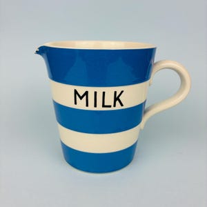 May include: A ceramic milk jug featuring horizontal blue and white stripes. The word MILK is printed in black capital letters on the central white stripe. The jug has a curved handle on the right side and a small pouring spout on the left.