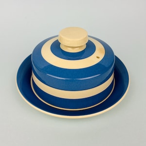May include: A ceramic butter dish featuring a circular base and a matching domed lid. The piece is decorated with alternating horizontal stripes of deep blue and cream. The lid is topped with a small, solid cream-colored knob handle.