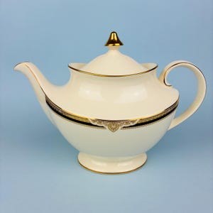 May include: A cream-coloured porcelain teapot featuring a gold-trimmed lid with a pointed finial. The body is decorated with a dark navy blue band accented by intricate gold filigree patterns along the centre and a gold-rimmed handle and spout.