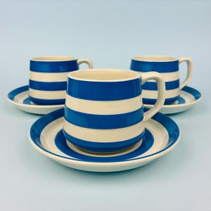 May include: A set of three ceramic cups and saucers featuring a horizontal striped pattern in vibrant blue and off-white. Each cup has a rounded body with a curved handle, and each saucer displays matching concentric blue and white stripes.
