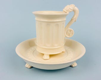 Taza y platillo de porcelana blanca sin marca, estilo gótico, con pie y asa de dragón.