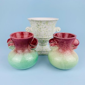 Könnte beinhalten: Drei Keramikvasen. Zwei kleine Vasen sind bordeauxrot und grün mit Henkeln. Die größere Vase ist weiß mit grünen Sprenkeln und Henkeln. Alle Vasen haben eine glänzende Oberfläche und stehen vor einem hellblauen Hintergrund.