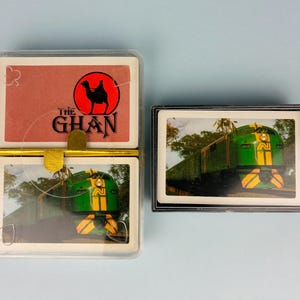 May include: Two clear plastic cases containing playing cards. The left case holds a deck with a red back featuring a black camel silhouette and the text THE GHAN, and a front card showing a green and yellow train. The right case shows a green train.