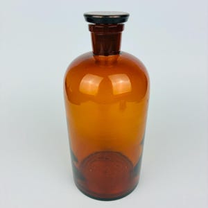 May include: A cylindrical amber glass apothecary bottle with a wide body and a narrow neck. The bottle features a thick, flat, black glass stopper that sits securely in the opening. The glass is translucent, allowing light to pass through.