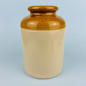 Könnte beinhalten: Ein Vintage-Steinzeugtopf mit zylindrischer Form. Der Topf hat einen hellbeige Körper mit braunem Deckel und Rand. Der Text "2pt" ist oben sichtbar. Der Topf wird wahrscheinlich zur Aufbewahrung verwendet und hat ein klassisches, rustikales Aussehen.